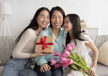 Loving multi generation Asian family with tulips and gift box hugging with closed eyes, celebrating Mother's Day at home. Senior woman with her daughter and granddaughter spending holiday together