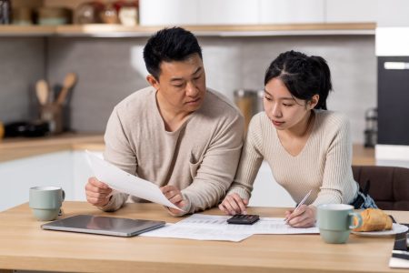 Serious asian family husband and wife sitting at kitchen table full of papers, laptop, coffee mugs, counting monthly expenses, using calculator, short of money. Economy, family budget concept