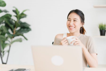 Beautiful young smiling asian woman working on laptop and drinking coffee in living room at home. Asia business woman working document finance and calculator in her home office. Enjoying time at home.